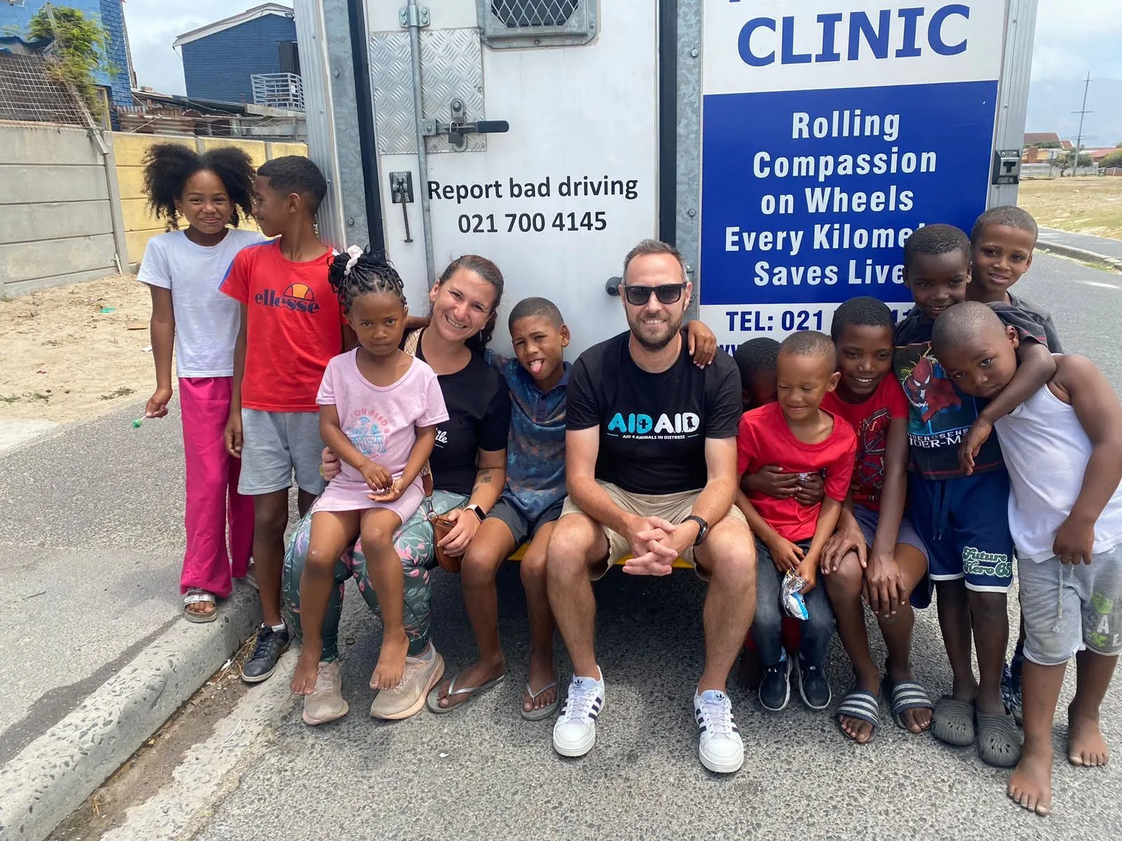 An Aid4Aid volunteer and a colleague sitting on the step of the mobile clinic with a group of children gathered around them.